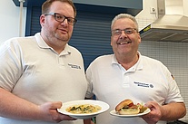 Zwei Männer in weißen Poloshirts stehen in einer Küche. Sie halten Teller, auf denen ein Gemüsecurry und ein Burger zu sehen sind.