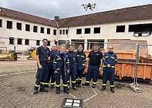 Eine Gruppen von Menschen in Einsatzkleidung des Technischen Hilfswerks steht vor einer Bauruine. Über ihnen schwebt eine Drohne.
