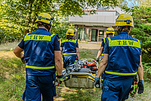 Vier Menschen in Einsatzkleidung des Technischen Hilfswerks tragen einen Schleifkorb auf ein Gebäude zu.