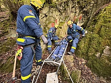 Einsatzkräfte stehen am Ausgang einer Höhle. Zwischen ihnen steht ein Metall-Schleifkorb zum Transport Verletzter.