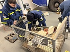 Einsatzkräfte beugen sich zu einem Stahlgitterkorb hinunter, in dem unter anderem Löschschläuche liegen.