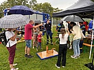 Mehrere Menschen stehen um eine Holzplatte herum. Einige von ihnen halten Schirme. 