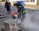 Zwei Menschen stehen auf dem Hof vor einem Gebäude. Einer hält einen Trennschleifer in den Händen und schneidet damit an einem Betonblock.